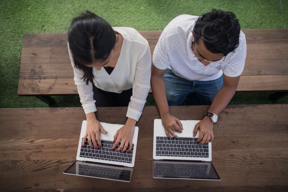 Overhead view of two colleagues working side by side on laptops at a wooden table